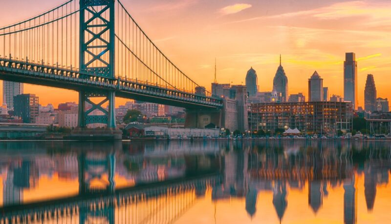 A romantic view of the Philadelphia skyline, water, and bridge at sunset.