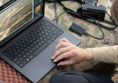 Lifestyle marketing photo of a person using a Windows laptop with a Samsung portable SSD plugged into it. A camera is also nearby, and it all sits on a green and orange surface.